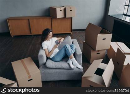 Girl having rest in new apartment and clicks phone. Successful independent spanish woman is moving. Living room interior and cardboard boxes. Mortgage loan and dream house purchase concept.. Girl having rest in new apartment and clicks phone. Independent spanish woman is moving.
