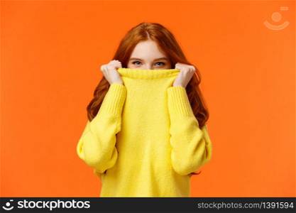 Girl hiding from winter cold in her warm soft sweater. Cute redhead woman pulling collar on nose and peeking at camera with smiling eyes, glancing at you happy, orange background.. Girl hiding from winter cold in her warm soft sweater. Cute redhead woman pulling collar on nose and peeking at camera with smiling eyes, glancing at you happy, orange background