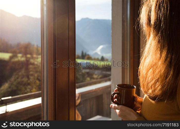 girl holding a cup of coffee looks out the window and mountains in the background