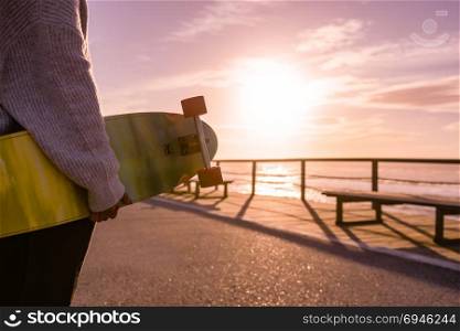 Girl holding a skateboard near the beach at sunset.