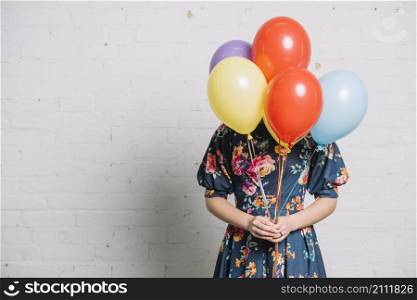 girl holding colorful balloons front her face standing against wall