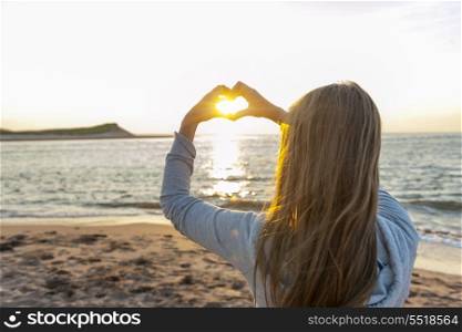 Girl holding hands in heart shape at beach. Blonde young girl holding hands in heart shape framing setting sun at sunset on ocean beach