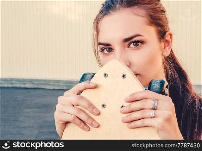 Girl holding skateboard in front of her face and looking at camera. A lot of space for text on wall behind the model.. Girl holding skateboard in front of her face and looking at camera.