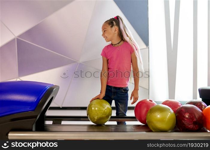 Girl holds a ball at the lane in bowling alley. Kid preparing to score a strike. Children having fun in entertainment center, little bowler. Girl holds a ball at the lane in bowling alley