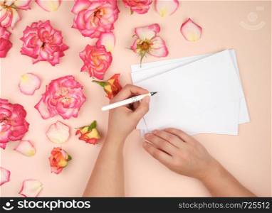 girl holds in her left hand a white pen and signs envelopes on a peach background with buds of blooming roses, top view