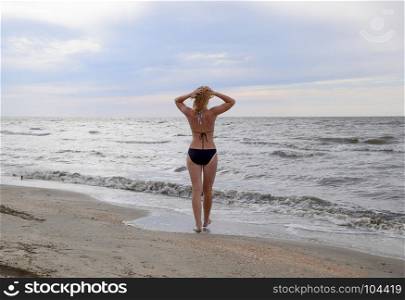 Girl in a bikini stands by the sea, a rear view. A beach walk of the blonde along the shore. The girl goes to the sea.. Girl in a bikini stands by the sea, a rear view. A beach walk of the blonde along the shore. The girl goes to the sea