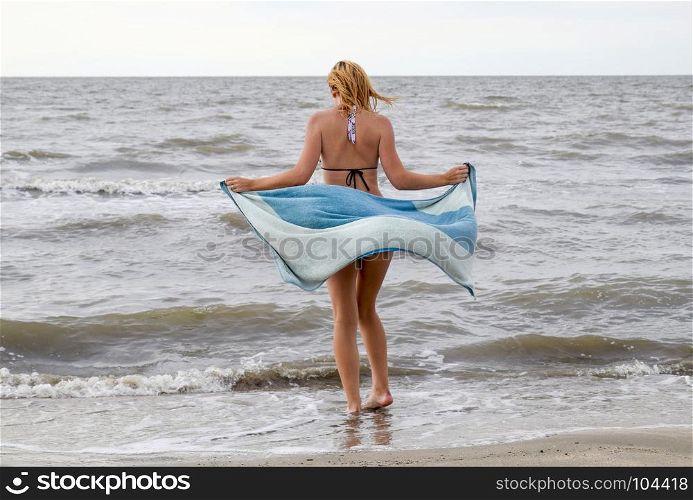 girl in a bikini with a towel in her hands is standing by the sea. A beach walk of the blonde along the shore.. girl in a bikini with a towel in her hands is standing by the sea. A beach walk of the blonde along the shore