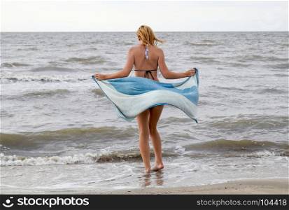 girl in a bikini with a towel in her hands is standing by the sea. A beach walk of the blonde along the shore.. girl in a bikini with a towel in her hands is standing by the sea. A beach walk of the blonde along the shore
