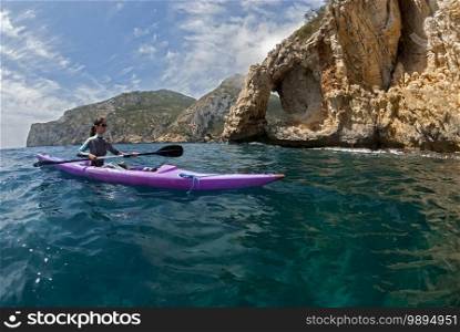 Girl in a kayak paddling close to a cliff with a big arch in the Mediterranean sea.
