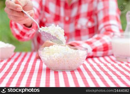 Girl in a plaid shirt picks up curd or cheese from a glass bowl with a spoon. There are dairy products on the table. Girl in plaid shirt picks up curd or cheese from glass bowl with spoon