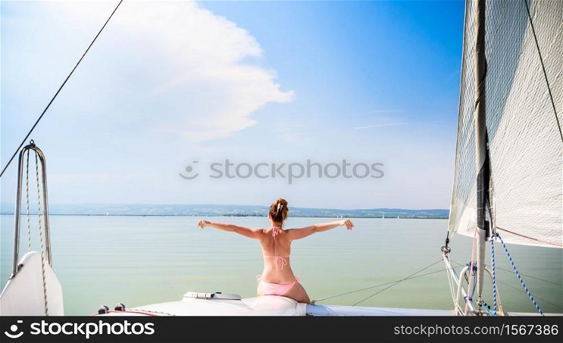Girl in bikini on a catamaran boat sits at front with hands spreed. Neusiedlersee lake in Austria Burgenland. Tourist spot destination for summer vacations. Girl on catamaran boat swims through Neusiedlersee lake