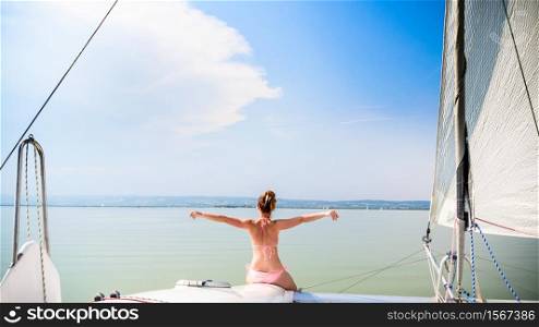 Girl in bikini on a catamaran boat sits at front with hands spreed. Neusiedlersee lake in Austria Burgenland. Tourist spot destination for summer vacations. Girl on catamaran boat swims through Neusiedlersee lake
