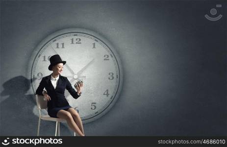 Girl in black cylinder. Pretty girl wearing retro hat siting on chair with clock in hand