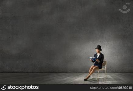 Girl in black cylinder. Pretty girl wearing retro hat sitting on chair with book in hands