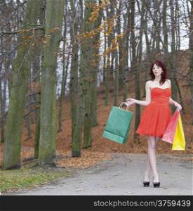 Girl in elegant red dress with colorful paper shopping bags walking in the autumnal park. Sale and retail.