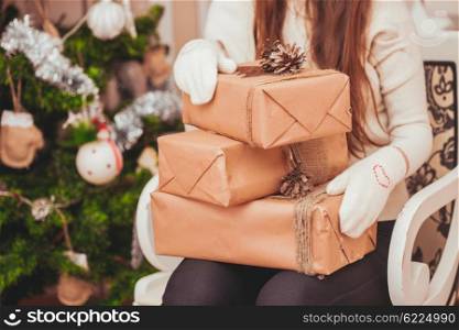 Girl in gloves holding gift boxes on her knees near Christmas tree. Girl with gift boxes