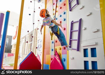 Girl in helmet is hanging on rope at the climbing wall, entertainment center, young climber. Children having fun, kids spend the weekend on playground, happy childhood. Girl is hanging on rope at the climbing wall