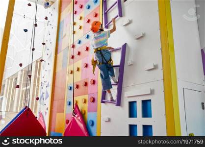 Girl in helmet is hanging on rope at the climbing wall, entertainment center, young climber. Children having fun, kids spend the weekend on playground, happy childhood. Girl is hanging on rope at the climbing wall