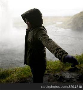 Girl in hooded coat standing in profile with arms spread before steamy body of water