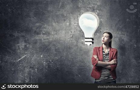 Girl in red jacket with arms crossed . Beautiful thoughtful girl looking up at bright light bulb on gray wall
