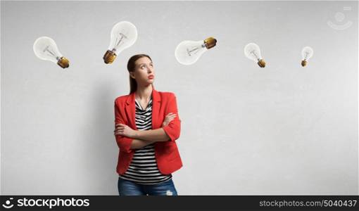 Girl in red jacket with arms crossed . Beautiful thoughtful girl looking up at bright light bulb on gray wall