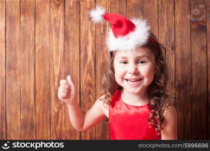 Girl in Santa hat headband is sitting over wooden wall and showing OK, copy space