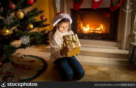 Girl in Santa hat sitting at burning fireplace and looking inside of Christmas gift box