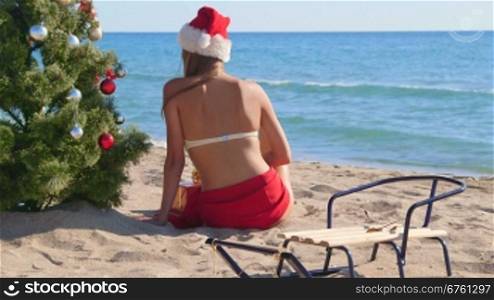 Girl in Santa hat sitting near Christmas tree on the beach rear view