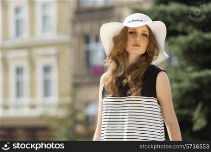 Girl in summer hat on the street. She is looking at camera and she wears striped dress and nice makeup.