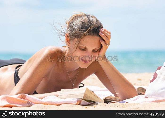 Girl in swimsuit reads book lying on sandy beach near the sea