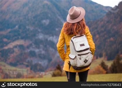 girl in the hat with a backpack stands on the meadow in the mountain