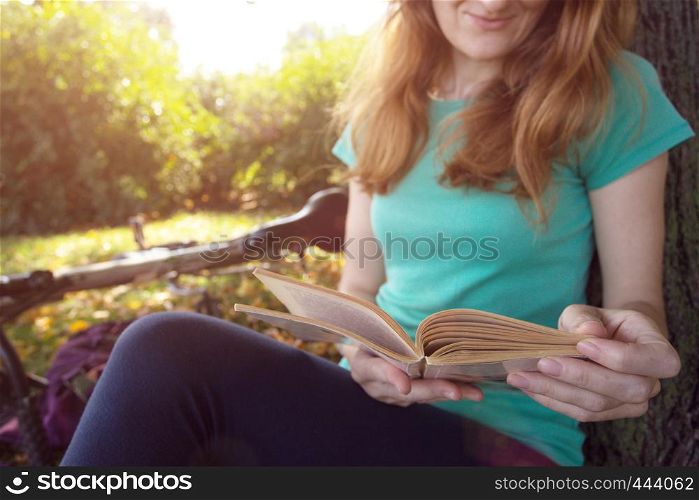 girl in the park sits under a tree and reads a book. next to it there is a bicycle and a backpack