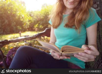 girl in the park sits under a tree and reads a book. next to it there is a bicycle and a backpack