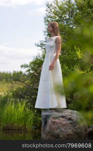 girl in white dress on the lake in the green against the sky stands on a stone