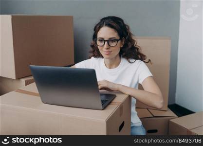 Girl in white t-shirt and glasses is texting on computer ordering delivery service. Happy hispanic woman packing boxes for moving to new place. Easy move concept.. Girl in white t-shirt and glasses is texting on computer ordering delivery service for packed boxes.