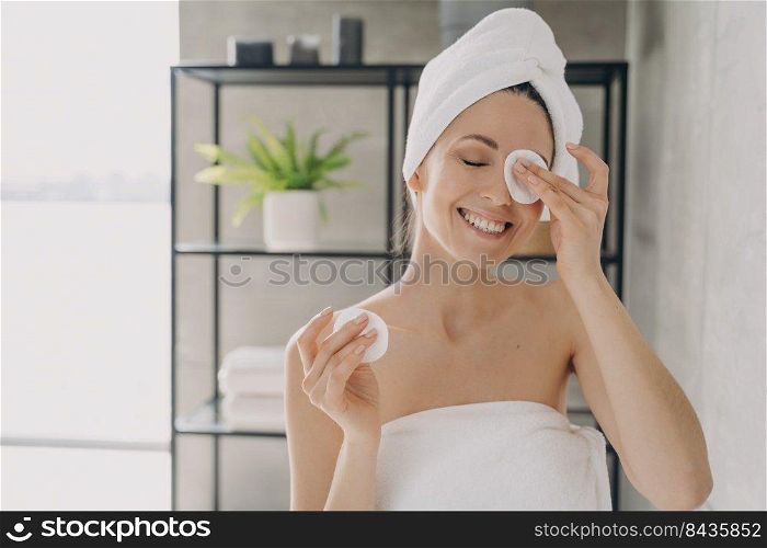 Girl is cleansing skin with face lotion in bathroom. Attractive caucasian woman wrapped in towel after bathing. Happy young woman takes shower at home. Delicate skin care and purifying.. Caucasian girl is cleansing skin with face lotion in bathroom. Delicate skin care and purifying.