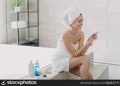 Girl is reading cosmetics ingredients list on the pack and smiling. Gorgeous european woman wrapped in towel after bathing. Young lady at spa resort. Concept of natural cosmetics and wellness.. Girl is reading cosmetics ingredients list on the pack and smiling. Concept of natural cosmetics.