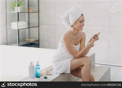 Girl is reading cosmetics ingredients list on the pack and smiling. Gorgeous european woman wrapped in towel after bathing. Young lady at spa resort. Concept of natural cosmetics and wellness.. Girl is reading cosmetics ingredients list on the pack and smiling. Concept of natural cosmetics.