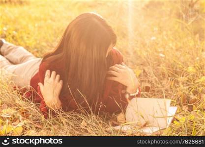 girl is reading in autumnal park, image backlit, she is looking at book and her face is hidden by her hair