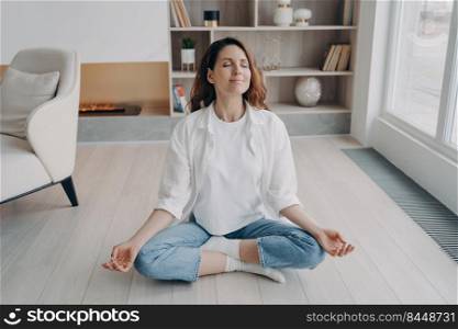 Girl is sitting in lotus pose with her eyes closed. Happy european woman is practicing yoga and meditation on floor at home. Morning gymnastics and zen. Stress relief, wellbeing and peace concept.. Girl is sitting in lotus pose with her eyes closed. Stress relief, wellbeing and peace concept.