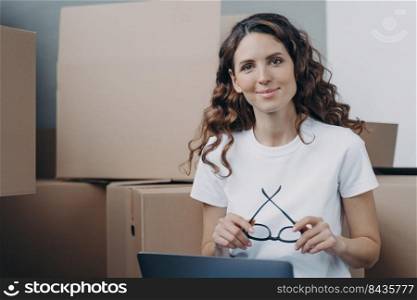 Girl is sitting on floor with laptop in new apartment. Happy hispanic woman in glasses with pc among cardboard boxes. Distance work from any place and freelance concept.. Hispanic girl is sitting on floor with laptop in new apartment among cardboard boxes.