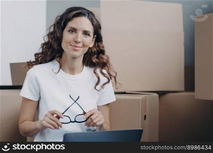 Girl is sitting on floor with laptop in new apartment. Happy hispanic woman in glasses with pc among cardboard boxes. Distance work from any place and freelance concept.. Hispanic girl is sitting on floor with laptop in new apartment among cardboard boxes.