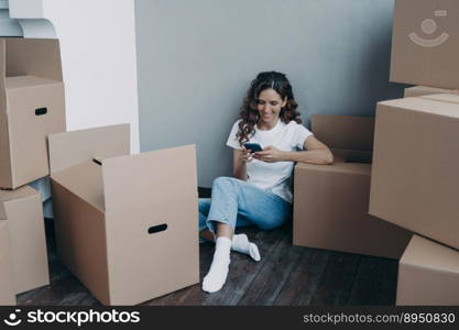 Girl is texting on mobile phone and sitting on the floor. Happy attractive spanish woman surrounded with cardboard packages and boxes. Easy moving to new apartment. Shipping service order concept.. Girl texting on mobile phone and sitting on the floor surrounded with cardboard packages and boxes.