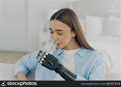 Girl is using futuristic robotic arm prosthesis and holding glass of water. Glass grip settings. European woman with artificial arm at home. Modern bionic prosthesis. Healthy lifestyle after surgery.. Girl is using futuristic arm prosthesis holding glass of water. Healthy lifestyle after&utation.