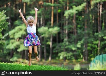 girl jumping on green lawn. Behind her is seen pine forest
