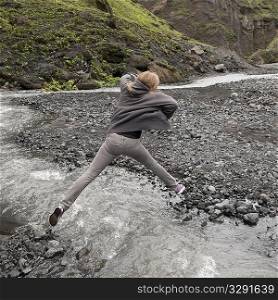 Girl leaping over fast flowing stream with rocky riverbank