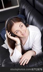 girl listening to music on her headphones at home lying on a black leather sofa