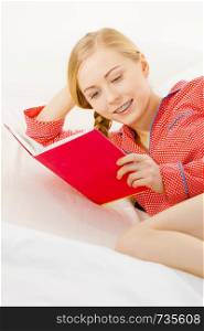 Girl lying in bed reading book. Young blonde female wearing red dotted pajamas relaxing at home on mattress.. Woman relaxing in bed reading book