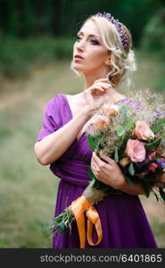 Girl model blonde in a lilac dress with a bouquet with a green forest