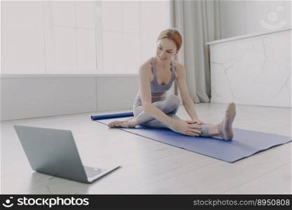 Girl on exercise mat practising stretching asana by online lesson. Young adult happy european woman enjoying yoga in living room. Concept of e-learning and home classes by computer.. Young adult happy woman enjoying yoga practising stretching asana by online lesson.